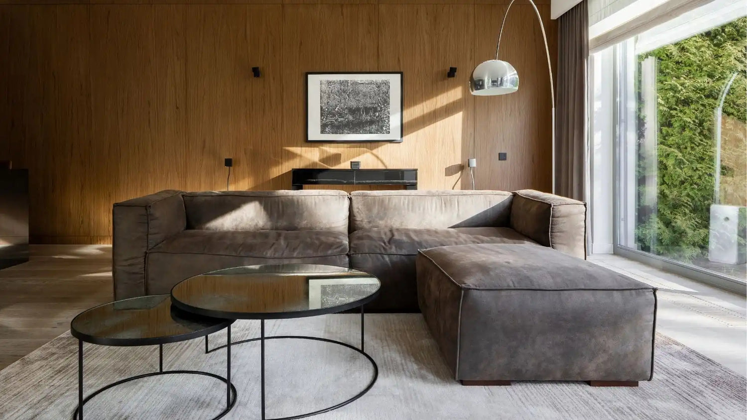 Un canapé sectionnel moderne, moelleux et gris, avec un pouf assorti et deux tables basses rondes en verre foncé avec des bases en métal noir.